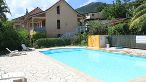 A swimming pool with lounge chairs, a stone-tiled deck, and a building with a balcony in the background. - Nice apartment in a quiet resort with pool only 50m from the beach. (Trois-Îlets)