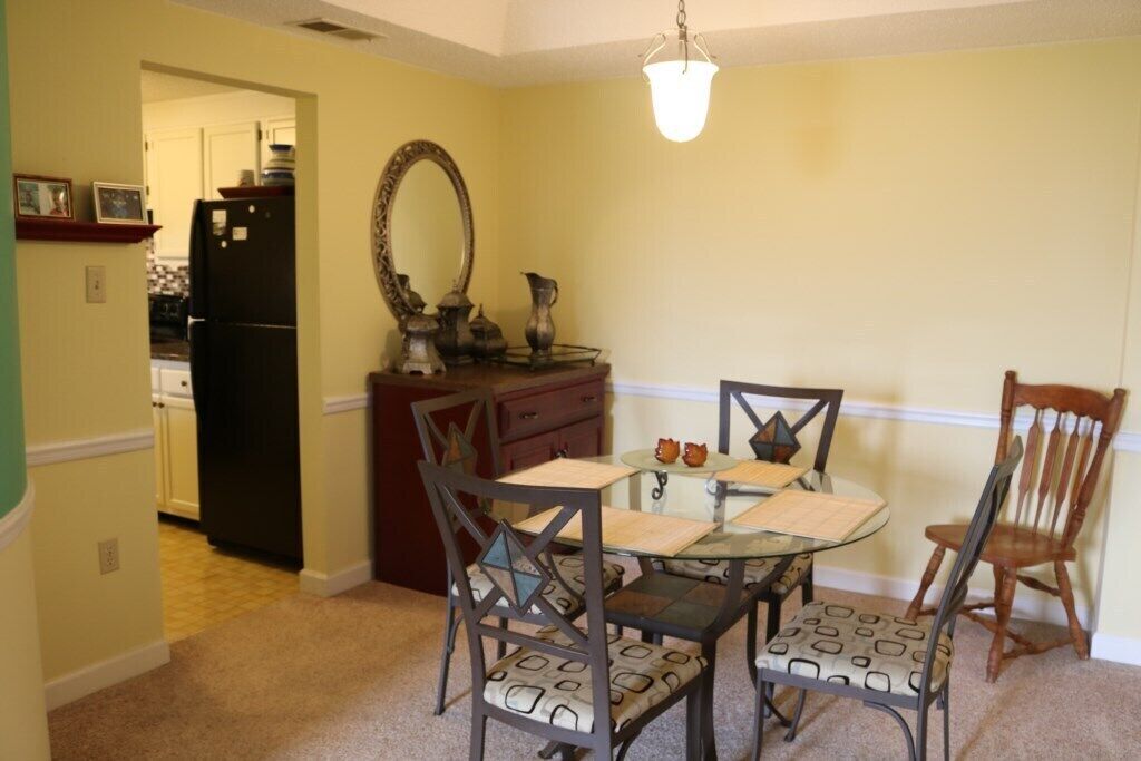 A dining area with a glass table, four chairs with patterned cushions, a wooden chair, a dark-colored refrigerator, a mirror, and a hanging light fixture.