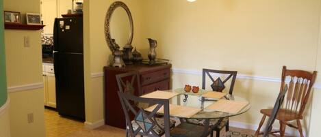 A dining area with a glass table, four chairs with patterned cushions, a wooden chair, a dark-colored refrigerator, a mirror, and a hanging light fixture.
