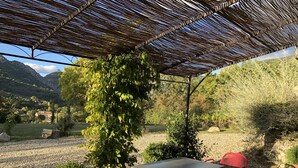 A covered outdoor seating area with a table and chairs, surrounded by greenery and a mountainous backdrop.