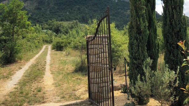 A gravel path leading to a metal gate, with cypress trees and a mountainous backdrop.