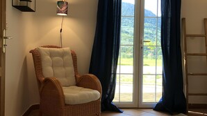 A room with a wicker chair, a small table, a window with curtains, and a view of greenery outside.