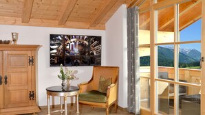 Ein Zimmer mit Holzbalkendecke, einem großen Fenster mit Bergblick, einem Holzschrank, einem runden Tisch mit Vase und einem grünen Polsterstuhl.