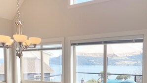A dining area with a wooden table and chairs, large windows with a view of a lake and mountains, and a chandelier. - Luxury Modern Lake Cottage, Huge Deck Sleeps 8, Pet Friendly, Close to Amenities (Kelowna)