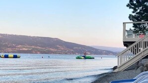 A beach with lounge chairs, a lifeguard tower, and floating barriers in the water. - Luxury Modern Lake Cottage, Huge Deck Sleeps 8, Pet Friendly, Close to Amenities (Kelowna)