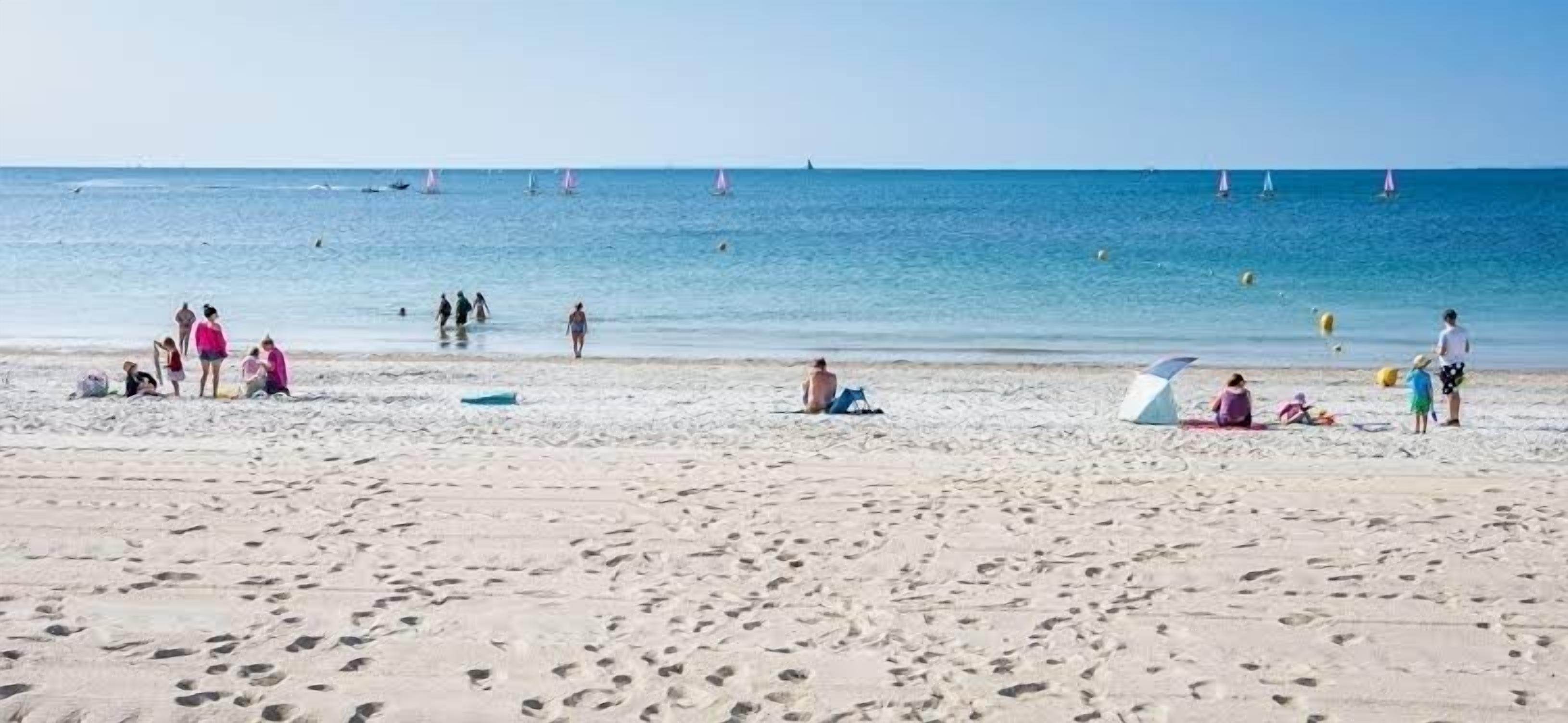Une plage de sable où se trouvent des gens, certains assis et d’autres debout, et quelques bateaux sur l’eau.