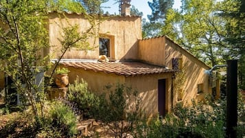 Un bâtiment rustique avec un toit en tuiles, entouré de verdure et agrémenté d’un banc en pierre.