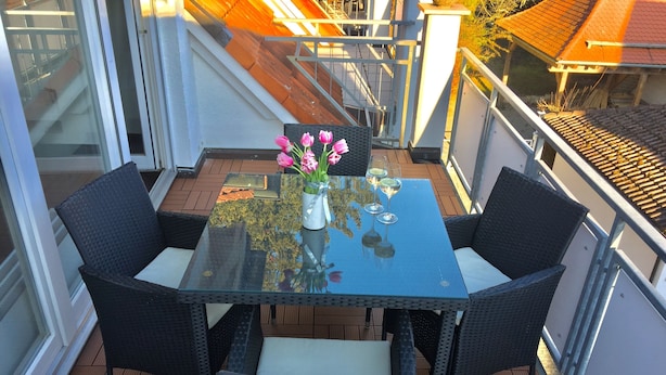 Una mesa con cubierta de vidrio, cuatro sillas de mimbre, un florero con flores rosadas y tres copas de vino sobre una terraza de madera.