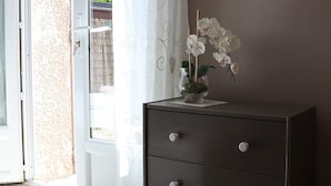 Une pièce dotée d’une porte-fenêtre coulissante donnant sur un espace extérieur, d’une commode avec des tiroirs, d’un miroir au mur et d’un vase avec des fleurs blanches posé sur la commode.