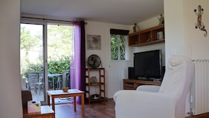 Un salon comprenant un fauteuil blanc, un téléviseur, une table en bois et une porte-fenêtre coulissante donnant sur un espace extérieur avec une table et des chaises de patio.