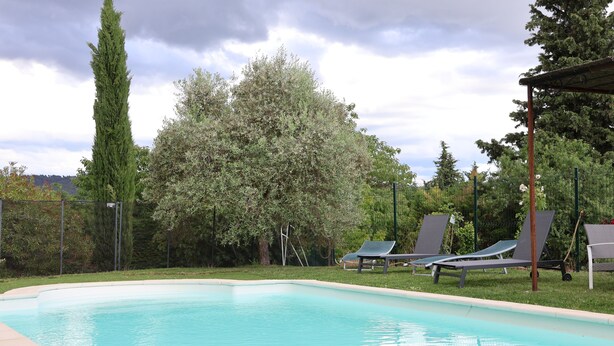 Une piscine à l’eau d’un bleu limpide, entourée d’une pelouse et d’arbres. On aperçoit des chaises longues et une pergola au fond.