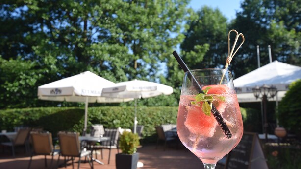 A glass of pink beverage with a mint garnish and a black chili pepper, set against an outdoor patio with tables and umbrellas. - Alte Mühle (Roedental)