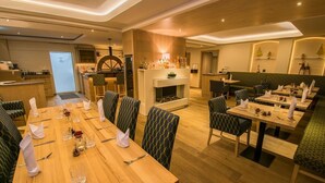 A modern dining area with wooden tables and green upholstered chairs, a fireplace, and a bar area with a wooden counter. - Alte Mühle (Roedental)