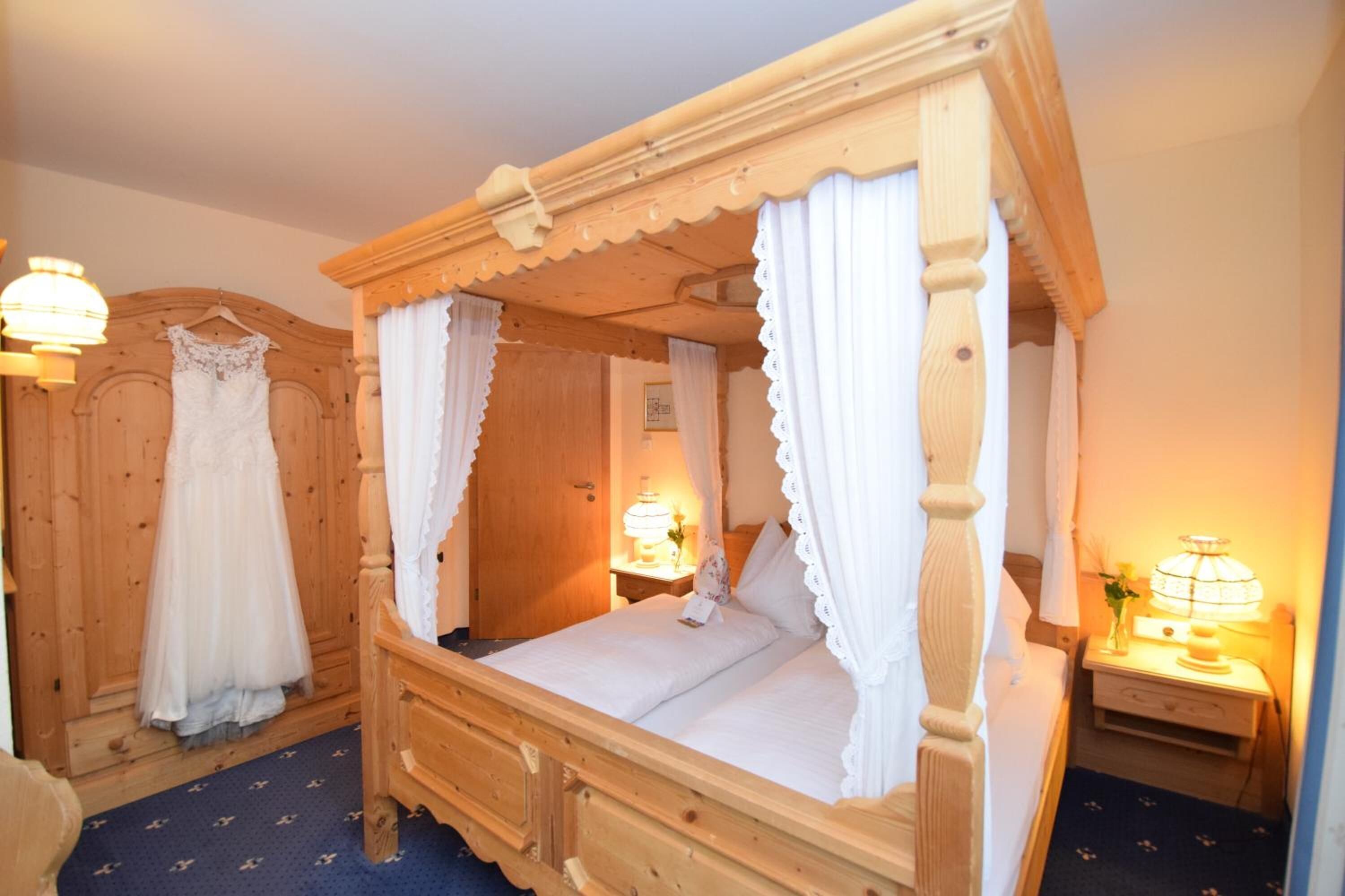 En seng med himmelseng i træ med hvide tekstiler og blonde detaljer, samt sengeborde med lamper.