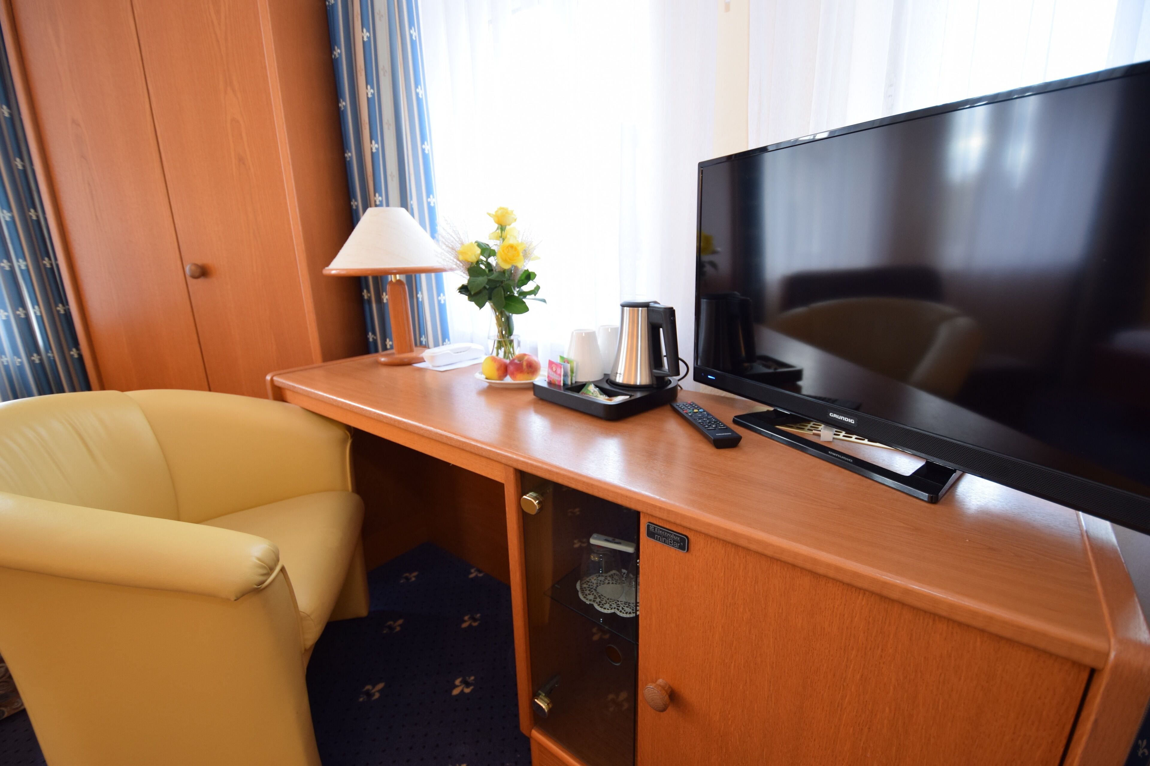 A hotel room with a wooden desk, a television, a lamp, a vase with flowers, and a chair.