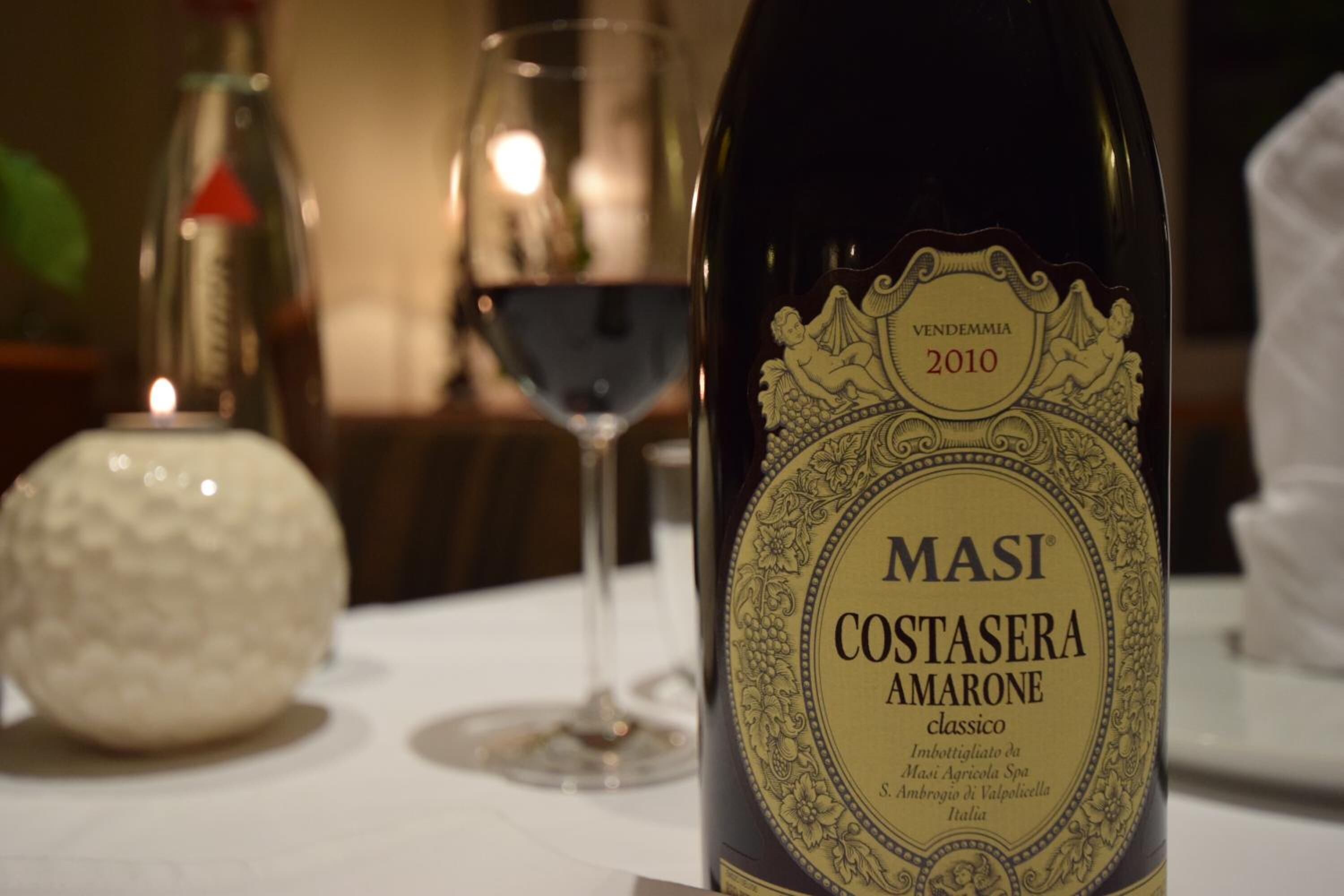 A bottle of Masi Costasera Amarone wine with a detailed label, placed on a table with a glass of red wine and a decorative object in the background.