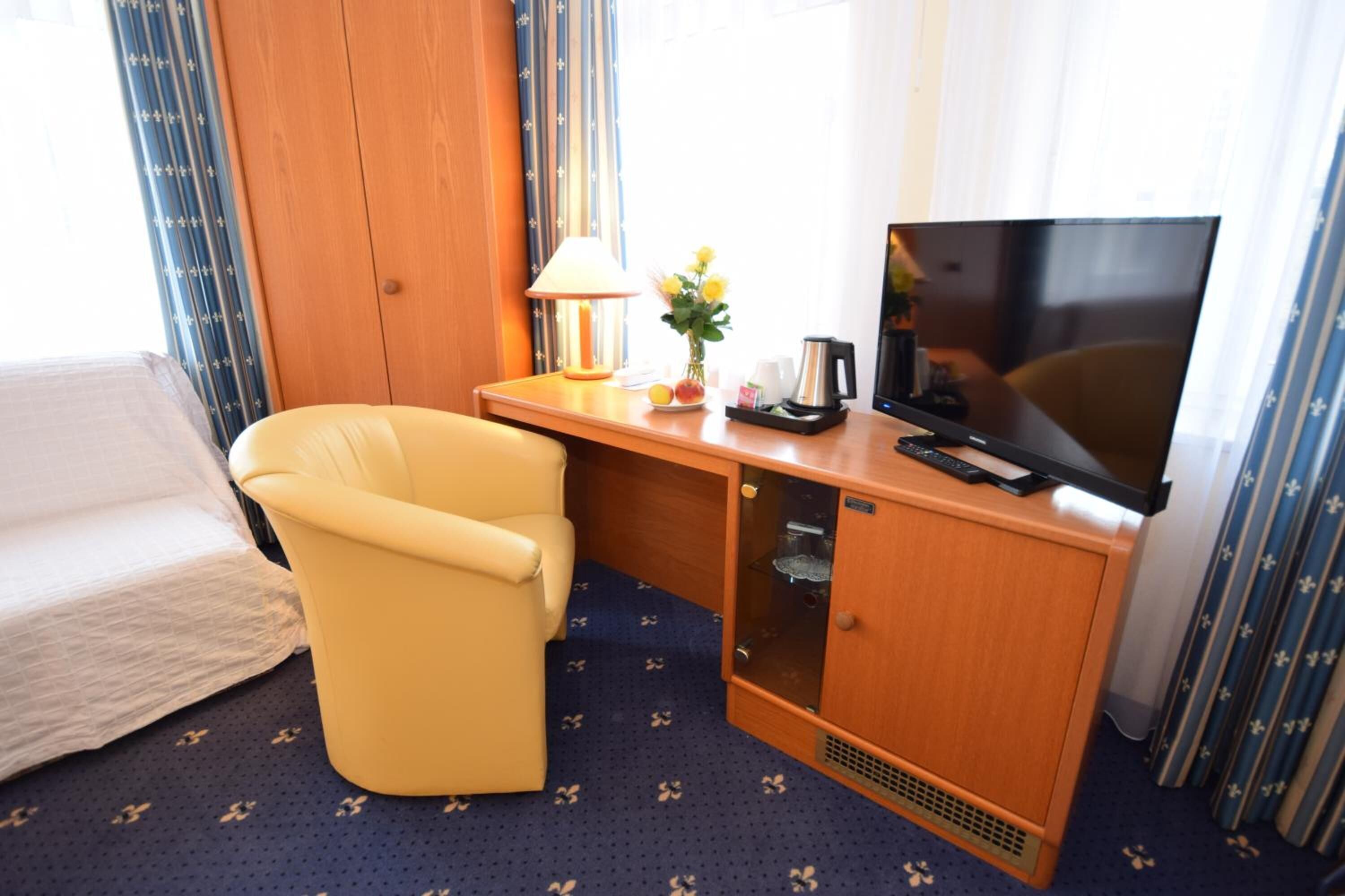 A hotel room with a bed, a yellow armchair, a wooden desk with a TV, a lamp, a vase with flowers, and a coffee maker.