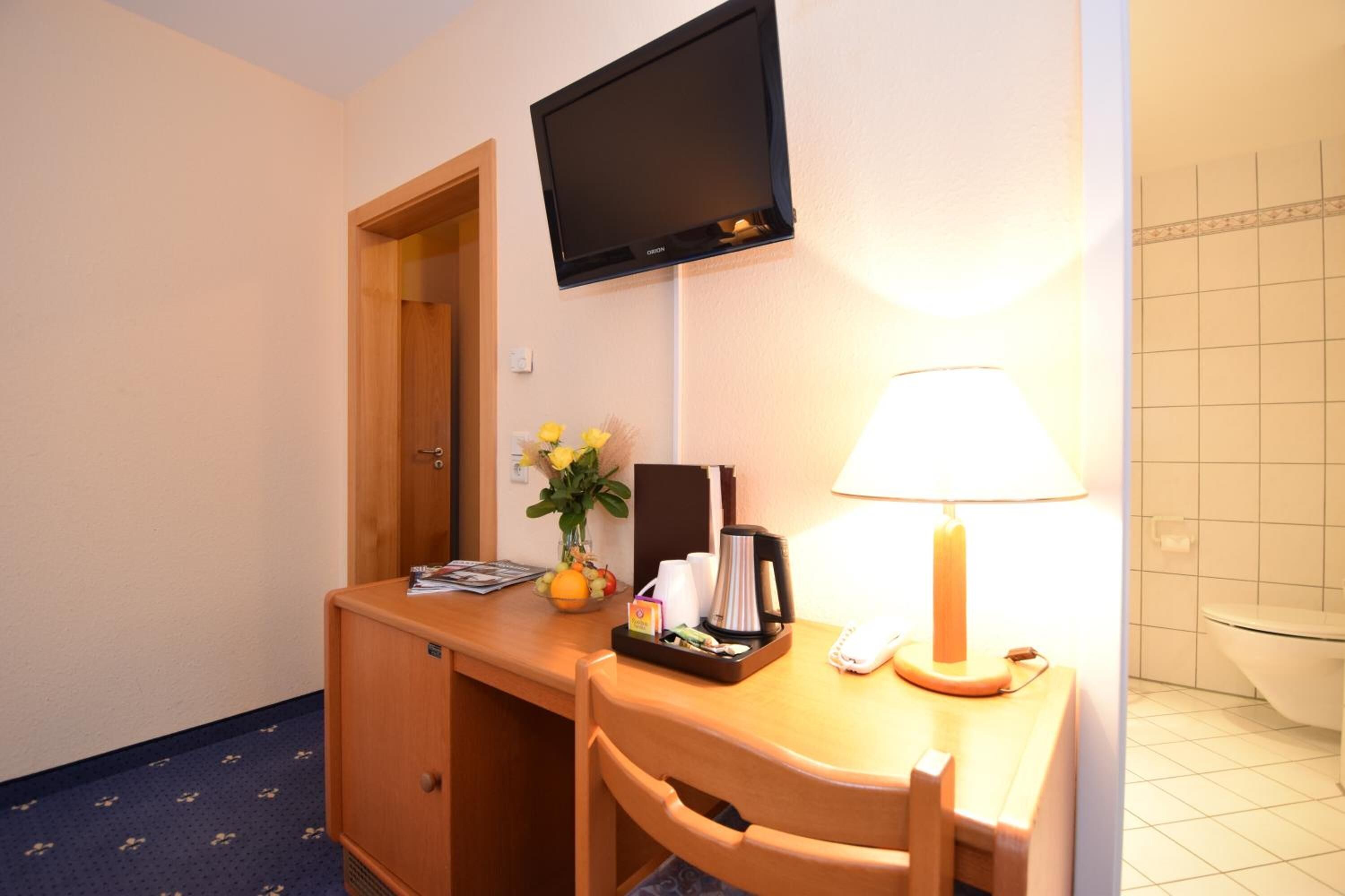 Habitación de hotel con escritorio, televisor de pantalla plana montado encima, lámpara, planta pequeña y baño con inodoro y paredes de azulejos.