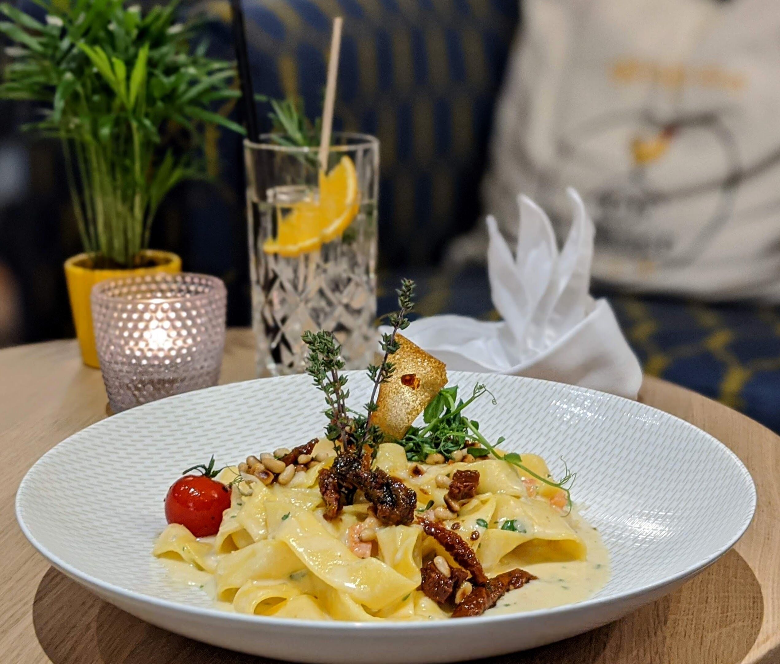 A plate of pasta with garnishes, a glass of water with a lemon slice, and a potted plant in the background.