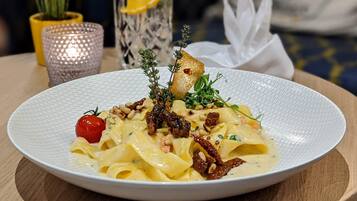 A plate of pasta with garnishes, a glass of water with a lemon slice, and a potted plant in the background.