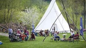 A group of people gathered around a white tent in a grassy area with trees in the background.