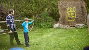 A person aiming a bow at a target with a crown symbol.