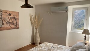 Un dormitorio con cama, aire acondicionado de pared, ventana con vistas a árboles, un cuadro de bicicleta y una alta jarra con plantas secas.