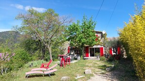 Un edificio de piedra rústico con contraventanas rojas, un jardín con sillas rojas y un cielo azul despejado.
