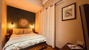Een slaapkamer met een bed, twee bedlampen, een wandspiegel, een ingelijst schilderij en een houten bankje.