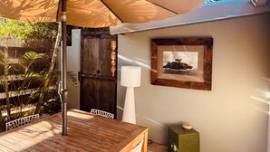 Een houten tafel onder een grote parasol, een witte stoel, een lijst met een foto aan de muur en een klein groen doosje.