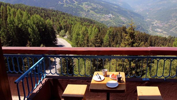 Un balcón con una mesa dispuesta para dos personas, con vistas a un paisaje montañoso que incluye una carretera y edificios en el valle.