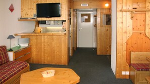 Habitación pequeña con interior de madera, televisor montado en la pared, zona de kitchenette, cama con cubrecama escocés y una mesita con una cesta.