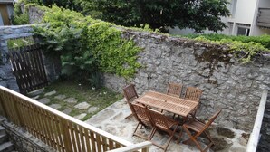 A stone patio with a wooden table and chairs, surrounded by a stone wall and greenery.