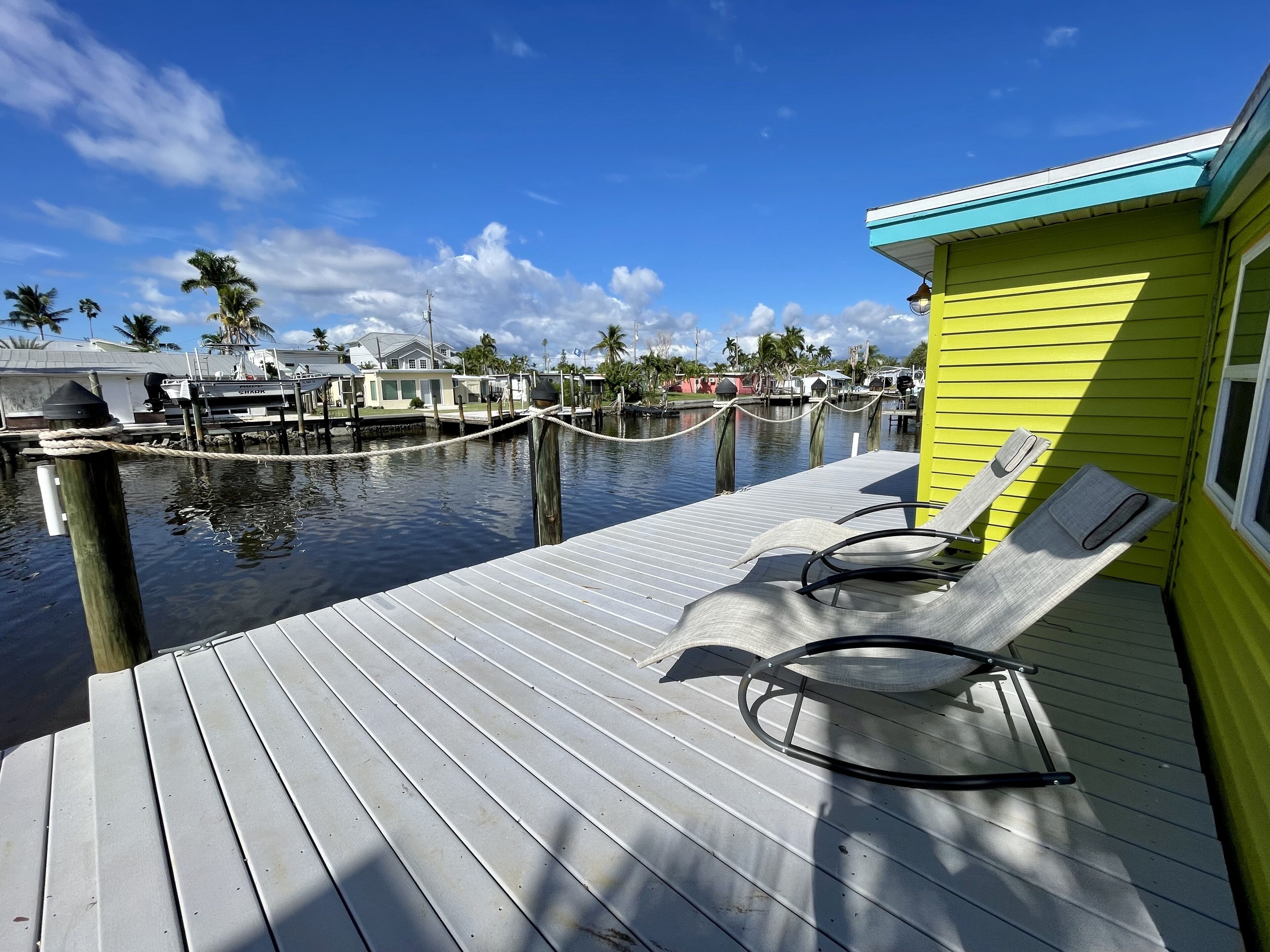 A wooden dock with lounge chairs, a yellow building, and a marina with boats.