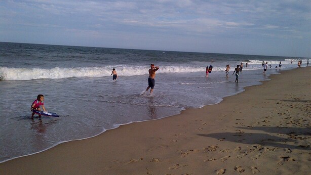 Una playa con gente caminando por la orilla y un niño jugando en el agua.