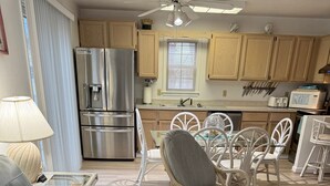 Una sala moderna con un sofá azul, sillas de mimbre, una mesa de centro de vidrio con conchas decorativas y una cocina con armarios de madera y electrodomésticos de acero inoxidable.