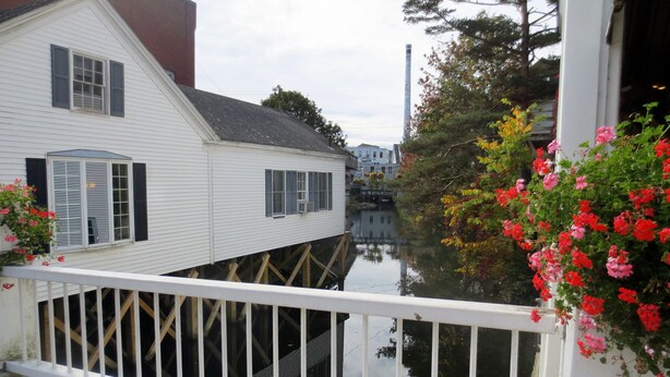 Een wit huis met een balkon dat uitkijkt over een gracht, houten steigers en een brug op de achtergrond.