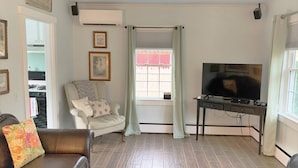 Een woonkamer met een bruine leren bank, een witte fauteuil met bloemenkussens, een televisie op een dressoir en een raam met gordijnen.