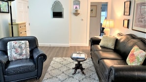 Een woonkamer met een zwarte leren bank, een zwarte fauteuil, een klein rond tafeltje en een wit vloerkleed.