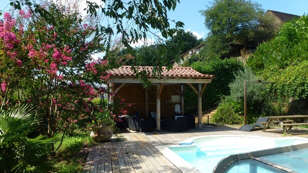 Une terrasse en bois avec une piscine, entourée d’une végétation luxuriante et de fleurs roses.