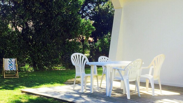 An outdoor patio with a white table and chairs, surrounded by greenery and a white wall.