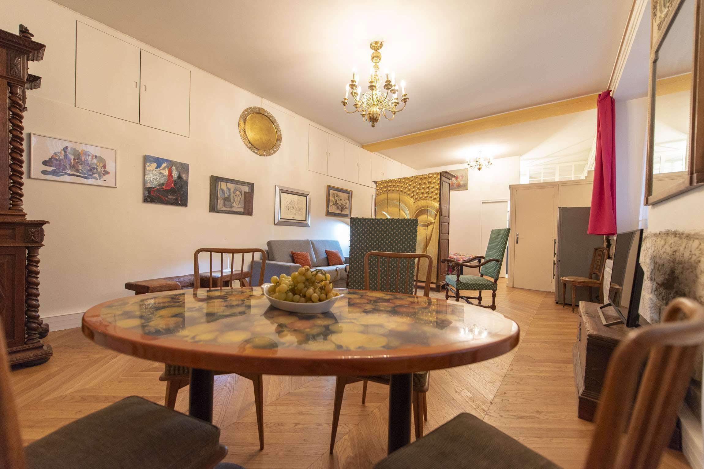 A dining area with a round table, chairs, and a bowl of fruit. There are framed pictures on the wall, a chandelier, and a patterned curtain.
