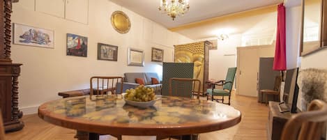 A dining area with a round table, chairs, and a bowl of fruit. There are framed pictures on the wall, a chandelier, and a patterned curtain.