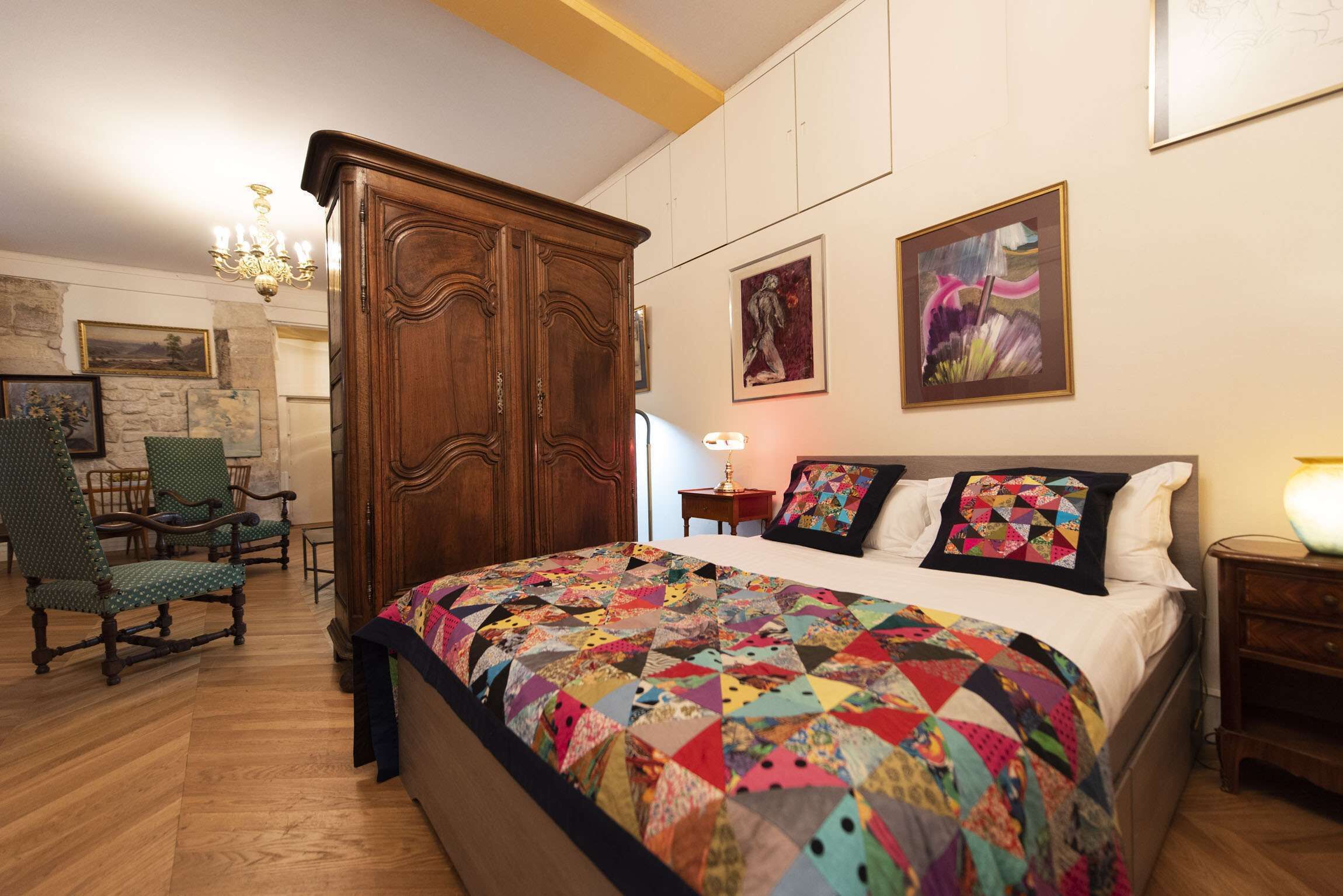 A bedroom with a large bed, wooden wardrobe, green upholstered chairs, and artwork on the walls.