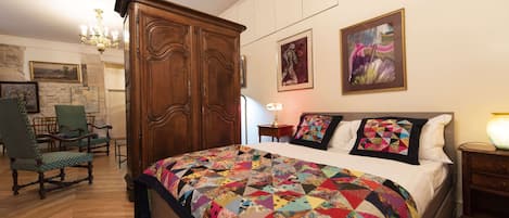 A bedroom with a large bed, wooden wardrobe, green upholstered chairs, and artwork on the walls.