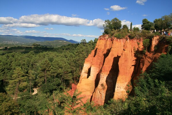 A natural landscape with a prominent red rock formation, dense green forest, and a distant view of mountains. - Completely renovated house (Oppède)