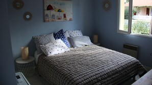 A bedroom with a patterned bedspread, a painting of umbrellas, and a window with a view of a building. - house 4/5 pers near lake and ocean (HOURTIN)