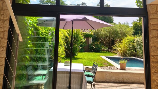Um pátio com mesa e cadeiras, um deck de madeira, piscina e vista para o jardim através de uma porta de vidro.