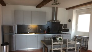 Et kompakt køkken med grå skabe, et rustfrit stål køleskab, indbygget mikrobølgeovn og gasblus. Der er et spisebord med fire stole og en skraldespand.