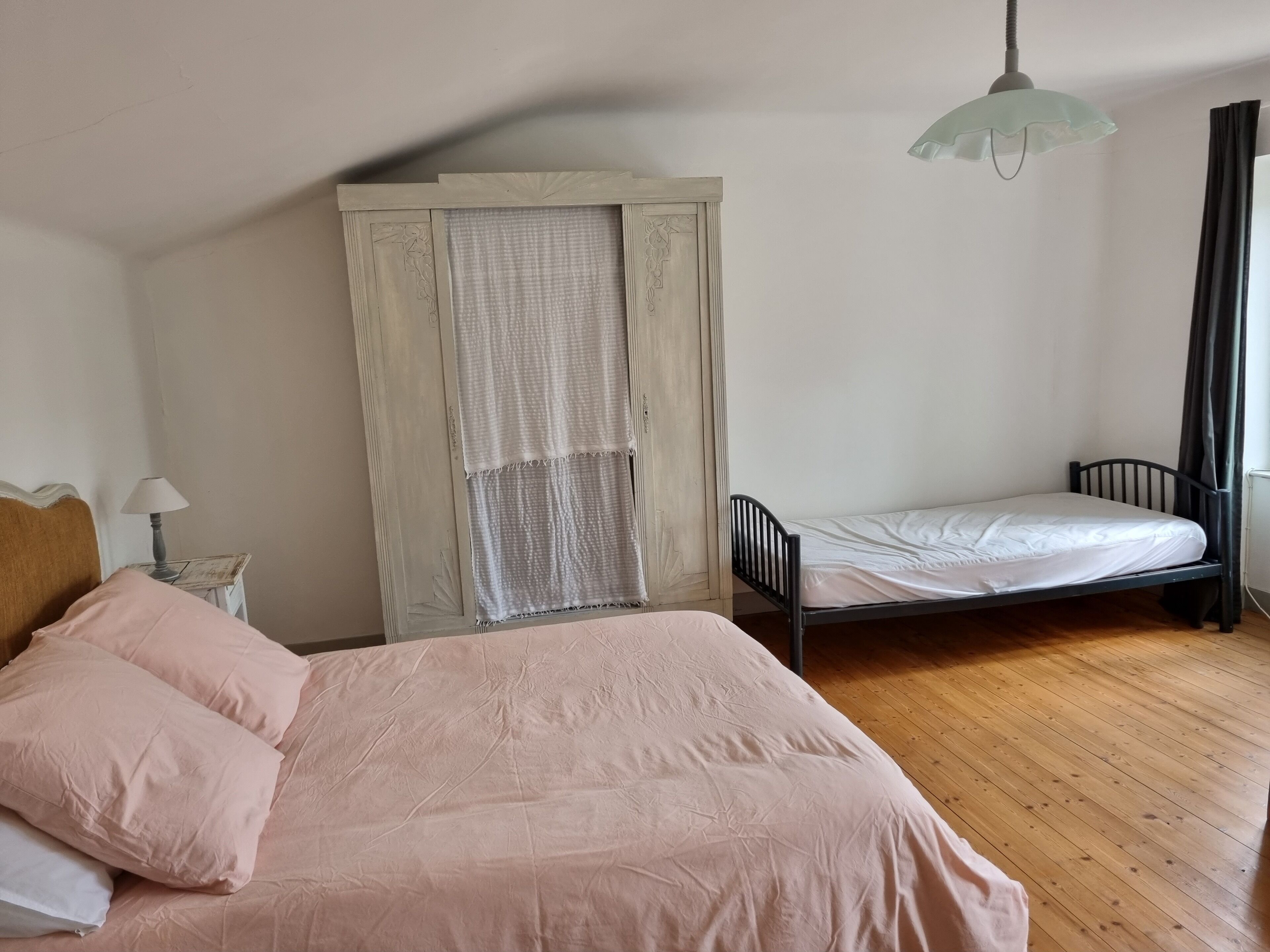 Une chambre avec un lit simple, une table de chevet, une lampe, une fenêtre avec des rideaux et un luminaire suspendu.